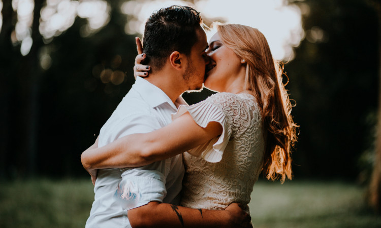 Casal de homem branco com camisa branca e mulher loira com camisola bege se beijando e sentindo apetite sexual excessivo.