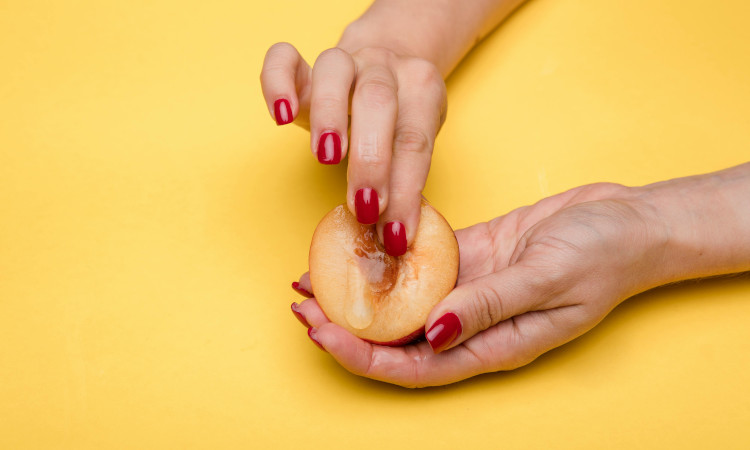 Mãos de mulher com unhas pintadas de vermelho mostrando como se dedar sozinha numa metade de um pêssego.