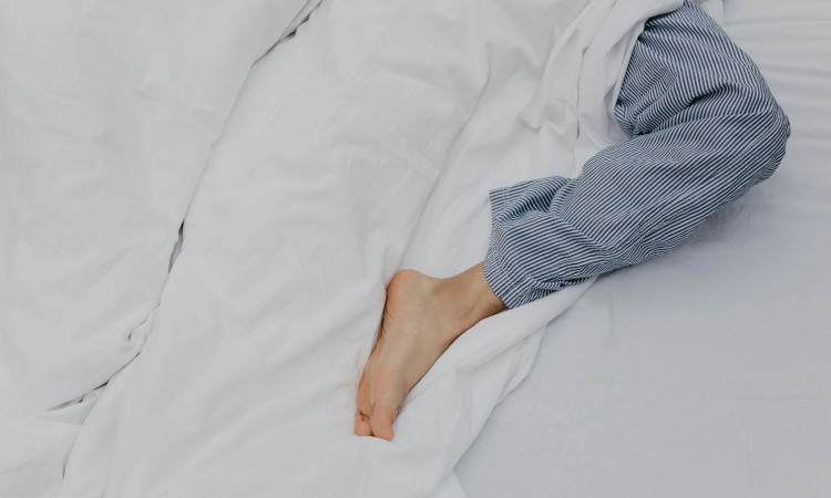 Perna de homem em pijama azul às riscas saindo debaixo de lençol branco após uma pessoa gozar dormindo.