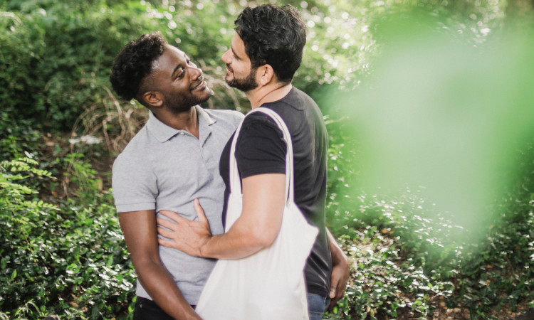Homem de pele negra, com camisa bege e sorrindo, pegando a bunda de homem de pele branca com barba e camisola preta buscando lugares públicos para fazer sexo gay.