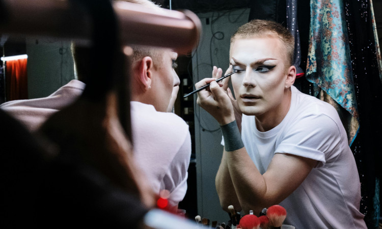Homem pintando os olhos e fazendo maquiagem de drag queen em frente a espelho.
