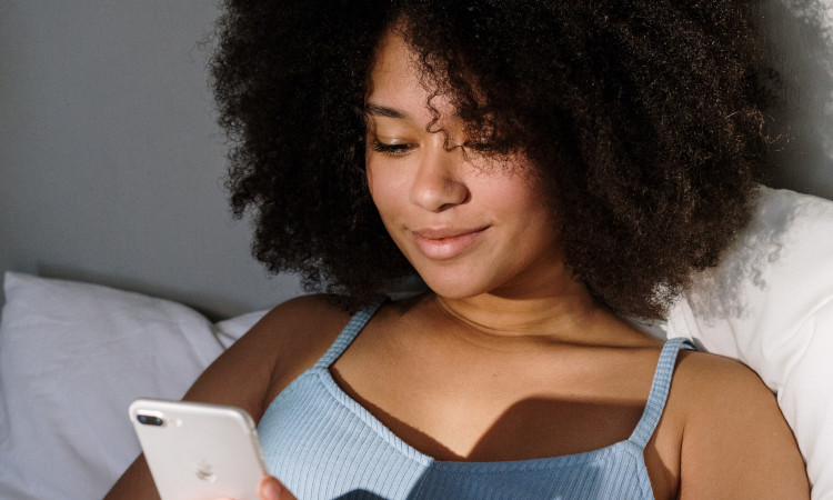 Jovem mulher morena e de cabelos afro castanhos olhando memes eróticos graciosos em seu celular deitada na cama.