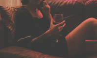 Mulher deitada num sofá, vestindo camisa de noite e segurando um copo de vinho na mão, falando mensagens safadas ao celular. 