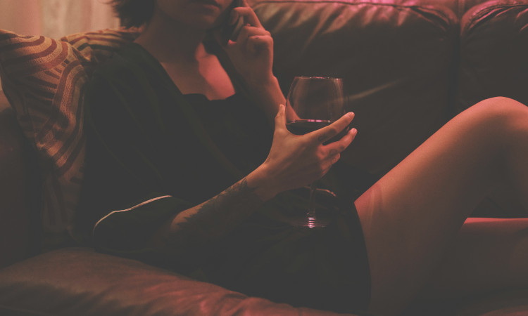Mulher deitada num sofá, vestindo camisa de noite e segurando um copo de vinho na mão, falando mensagens safadas ao celular. 