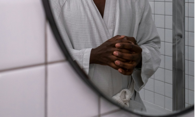 Tronco de homem negro, com mãos na frente e de roupão branco, se olhando ao espelho e pensando em o que é dismorfia sexual.