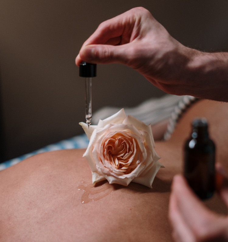 Mãos de homem com gotejador deitando fio de óleo nas costas de mulher, com uma flor rosa, antes de sexo com óleo de coco.