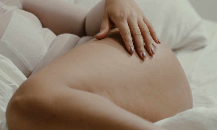 Coxa de mulher deitada na cama, vestindo lingerie branca e sendo acariciada com mão de unhas pintadas de um bege escuro pensando em como fazer sexo sozinha.