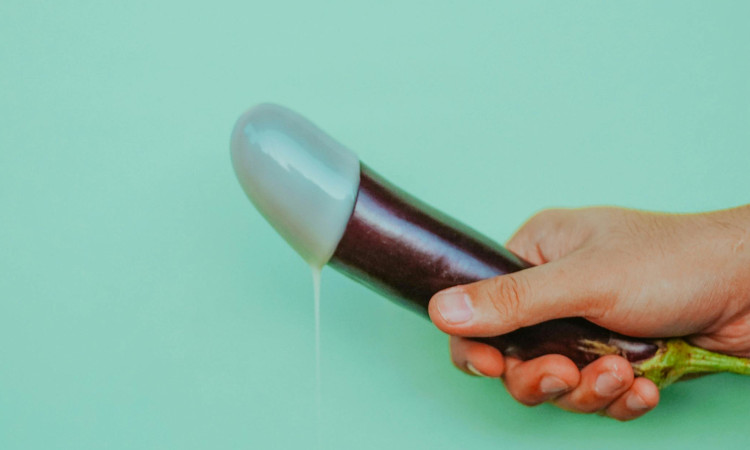 Mão de homem segurando beringela como se estivesse se masturbando numa imagem com fundo verde e uma substância branca imitando esperma escorrendo da ponta.